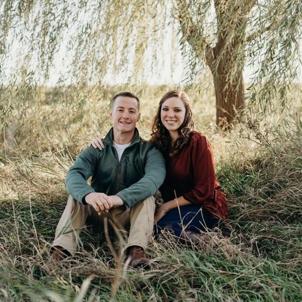 Matt Wood and wife sitting in a field under a tree. Matt is wearing a green zip-up with khaki pants, while the woman is wearing a burnt orange top. 
